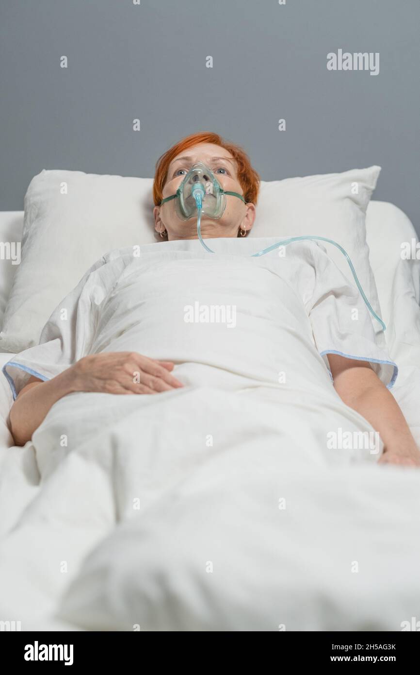 Donna anziana in maschera di ossigeno che si trova sul letto e respira con speciale apparecchiatura medica in ospedale reparto durante il coronavirus Foto Stock