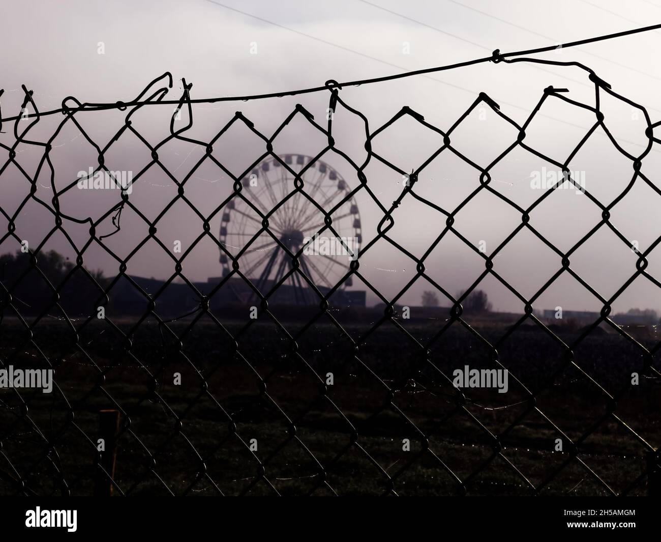 Recinzione di sicurezza filo spinato con grande campo e giostra ferris ruota merry andare rotondo in background visto attraverso nebbia Foto Stock