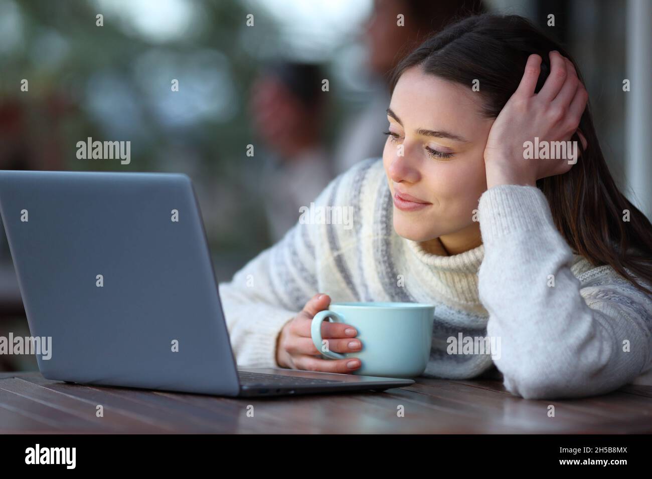 Donna felice che tiene la tazza di caffè che controlla il contenuto del laptop in inverno in una terrazza Foto Stock