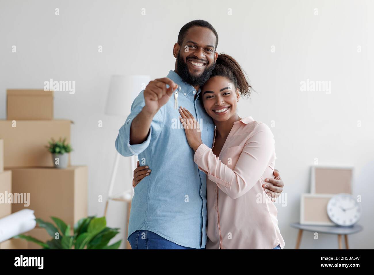 Felice sorridente giovane afroamericana amorevole famiglia abbracciando in nuova casa tra le scatole e mostrando le chiavi della casa Foto Stock
