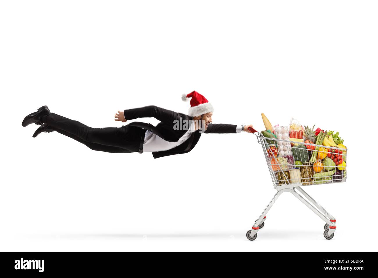 Scatto a tutta lunghezza di un uomo d'affari con un cappello di natale babbo natale che vola e tiene un carrello con cibo isolato su sfondo bianco Foto Stock