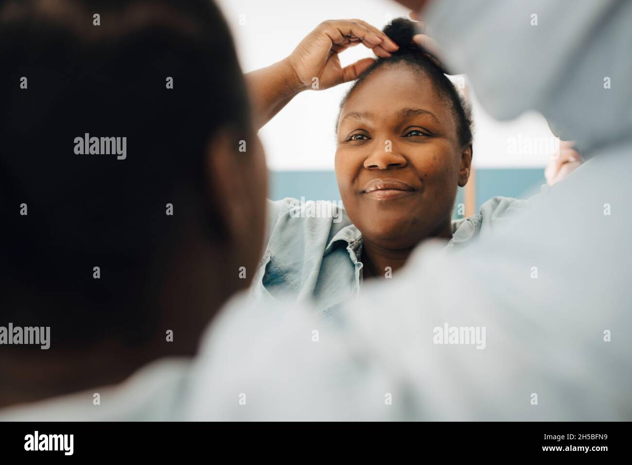 Donna sorridente che regola i capelli mentre guarda nello specchio a casa Foto Stock