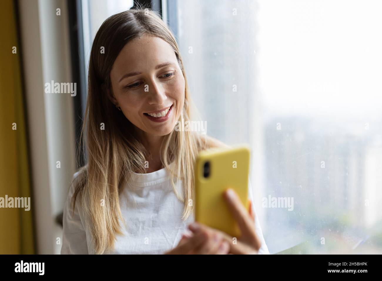 Giovane donna graziosa millenaria che tiene il telefono cellulare a casa o caffè vicino alla finestra. Mockup con spazio di copia Foto Stock