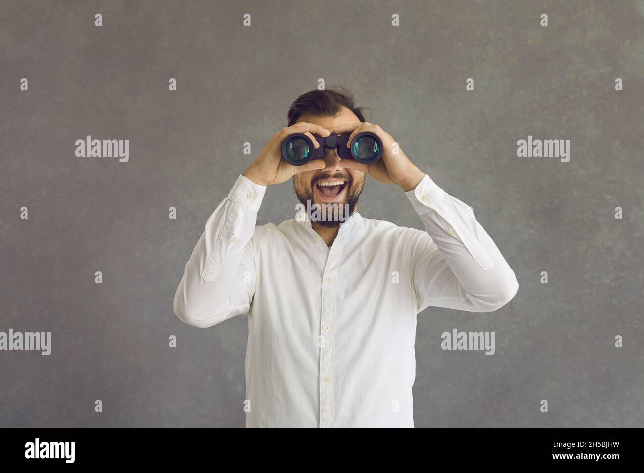 Giovane uomo che guarda attraverso binocoli su sfondo grigio studio con volto felice Foto Stock