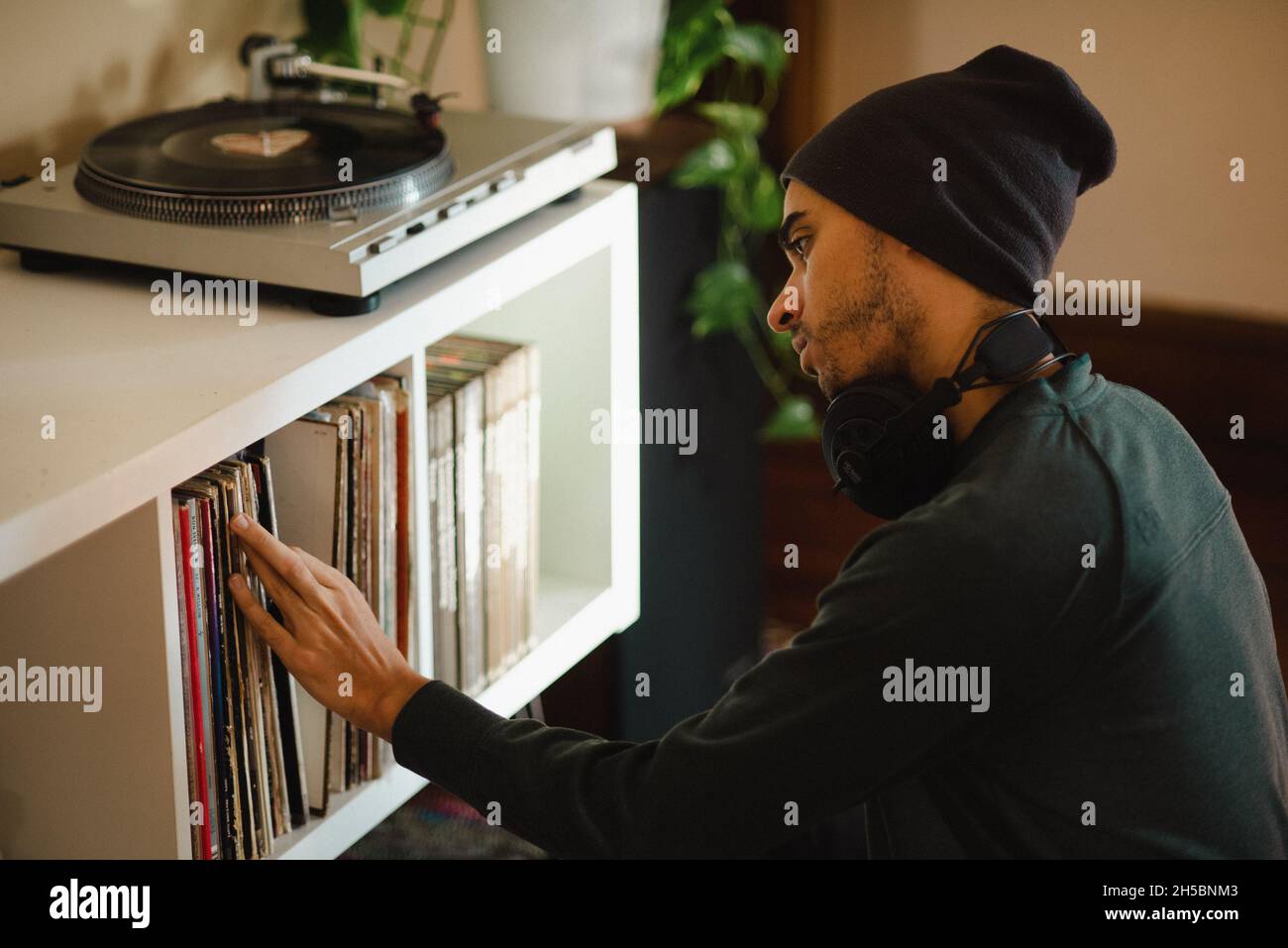 Giovane uomo da millennial mixed-race in beanie con cuffie che esplorano la collezione di dischi in vinile, che sta per ascoltare la musica. Foto Stock