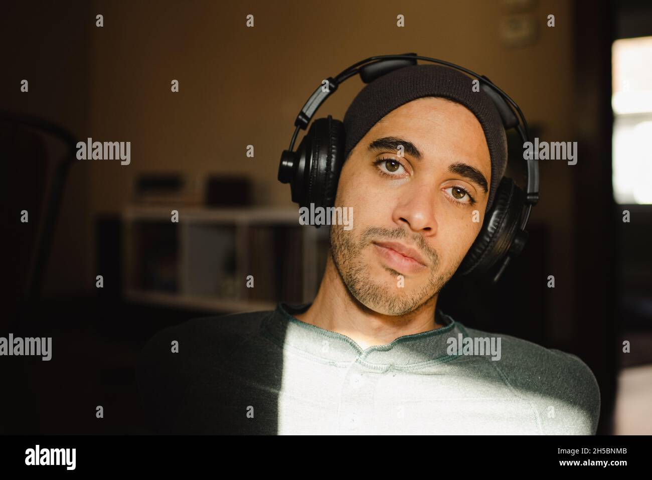 Giovane uomo millenario con un occhio di nocciola di razza mista che ascolta la musica con le cuffie sovrauricolari e guarda la fotocamera. Hipster con cuffie e beanie al sole Foto Stock