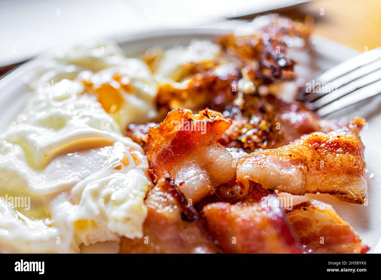 Macrofumo di strisce di pancetta fresca fritte fatte in casa e uova su piatto bianco con forchetta sul tavolo da cucina in legno Foto Stock