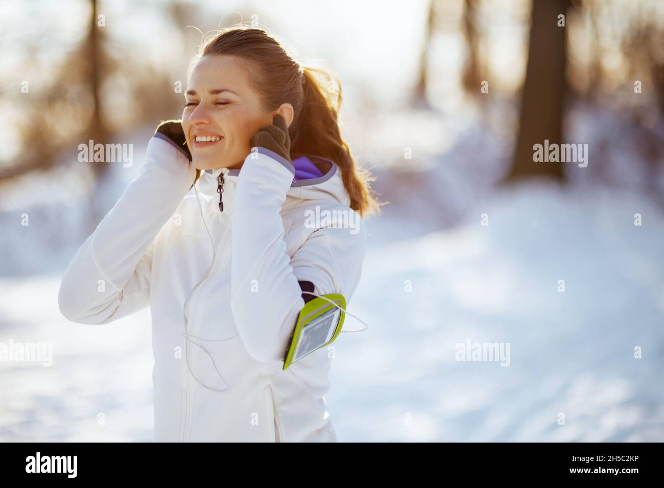 donna sana sorridente in giacca bianca con cuffie all'aperto nel parco cittadino in inverno. Foto Stock