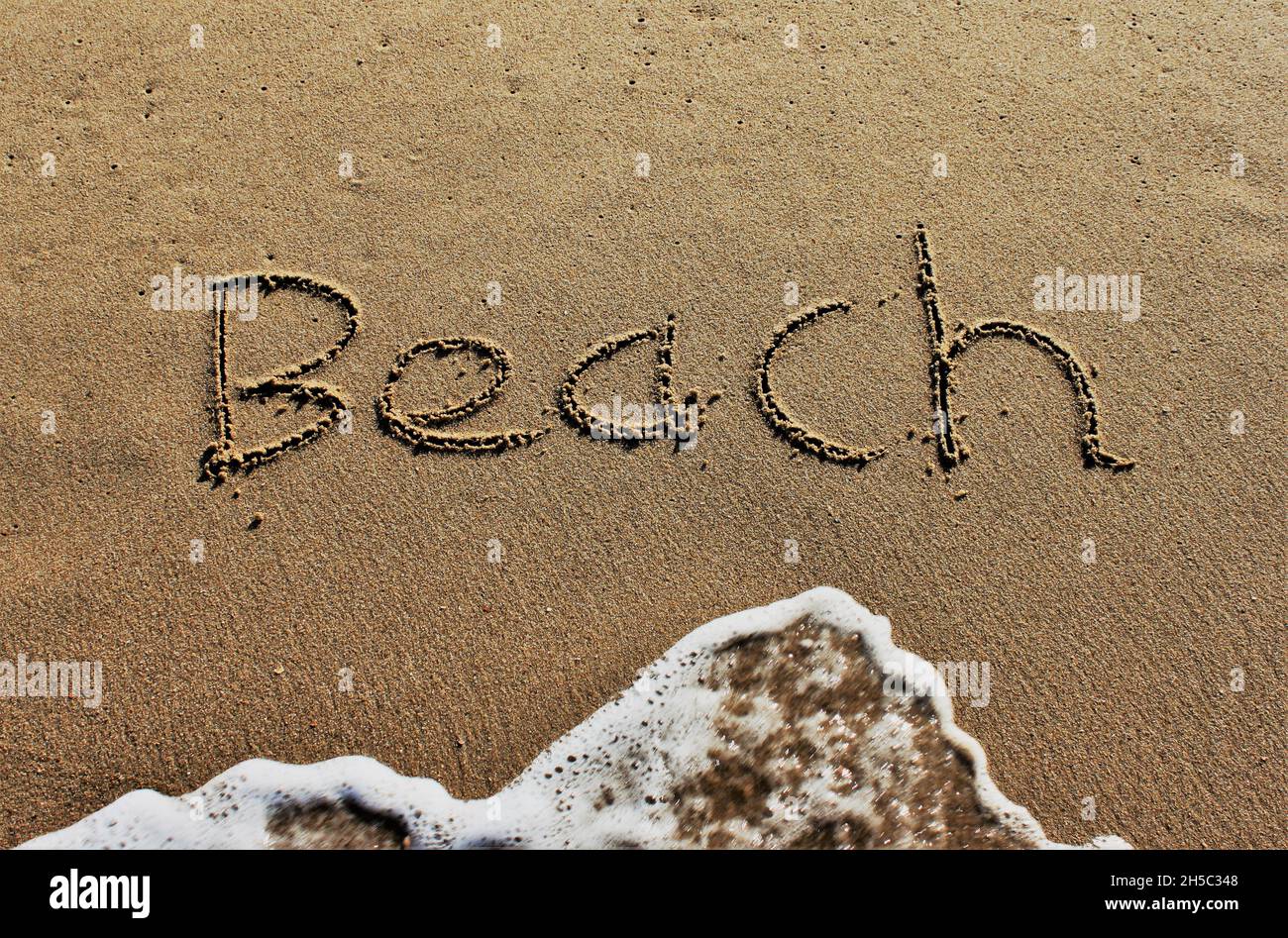 Sulla spiaggia, la parola Spiaggia è scolpita in lettere nella sabbia liscia, che è lavata via da un'onda Foto Stock