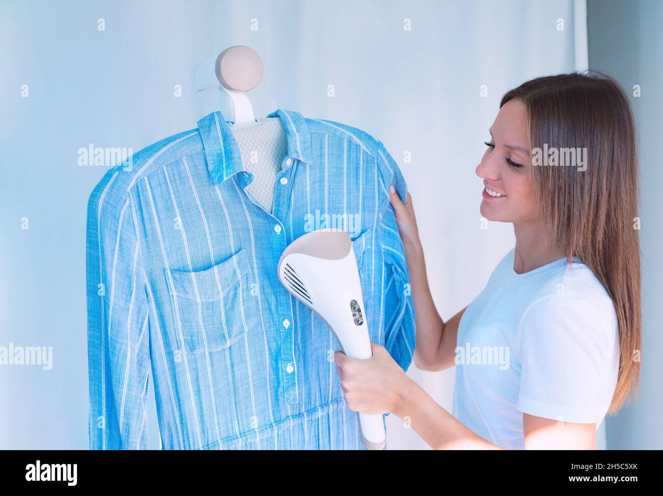 Donna abito blu da stiro con vaporetto per indumenti. Donna con vaporetto pulizia abiti a casa Foto Stock