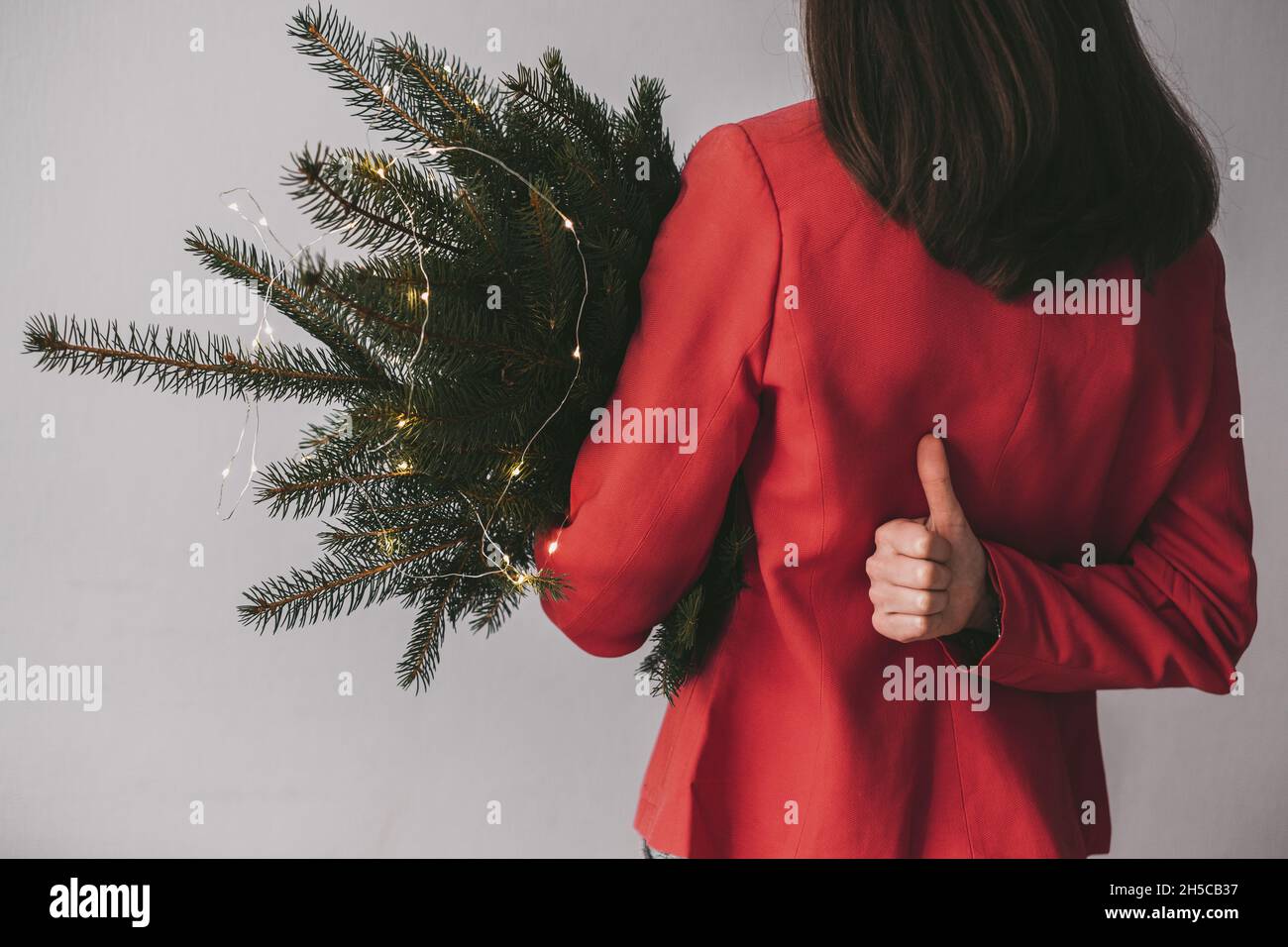 Ragazza girò la schiena mostrando il pollice in su in una giacca rossa che tiene un ramo dell'albero di Natale su uno sfondo grigio. concetto di conservazione della foresta. Alta qualità Foto Stock