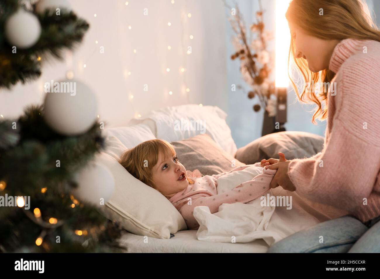 Madre mette sua figlia a letto in una stanza accogliente decorata per natale Foto Stock