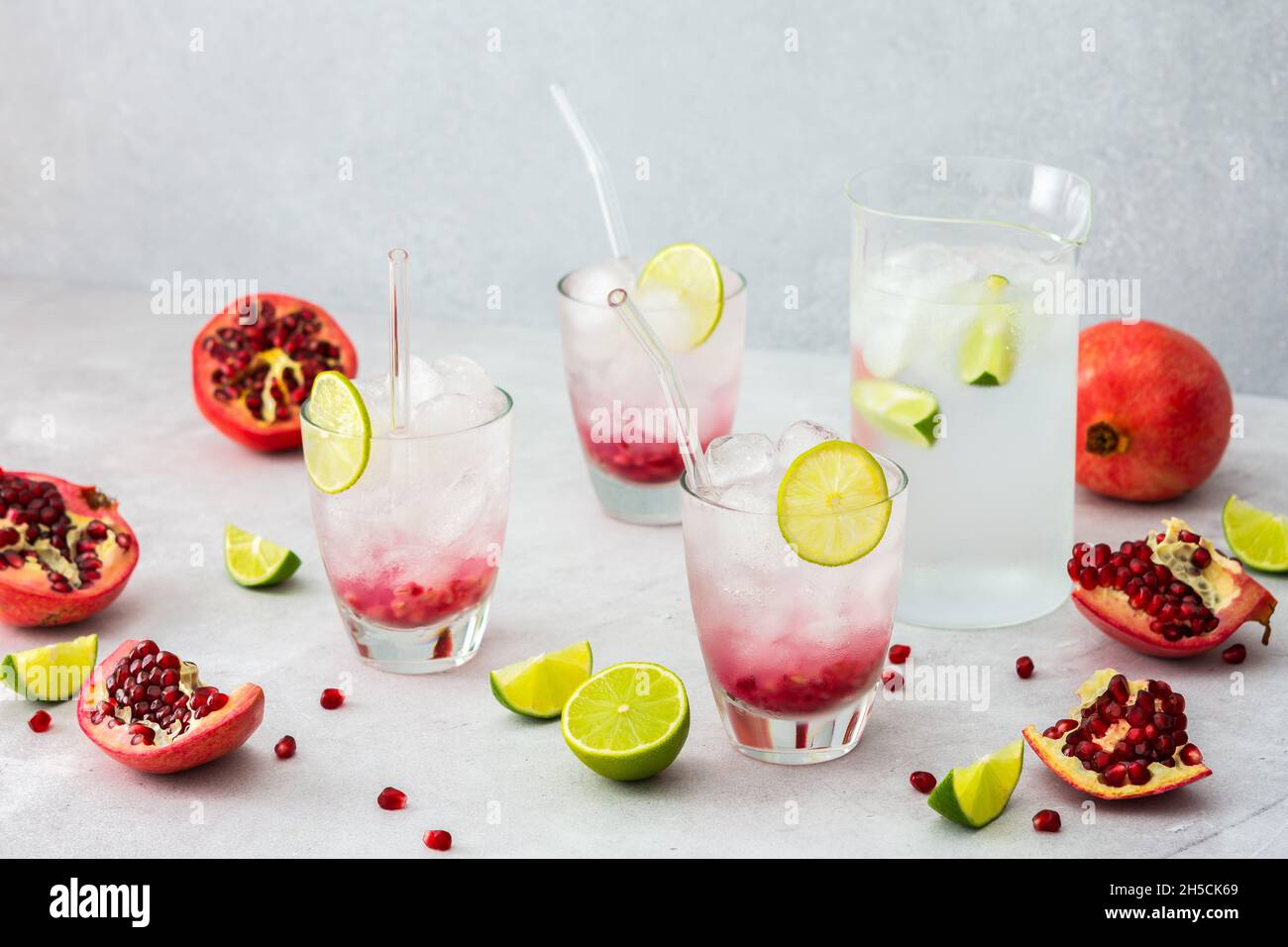 Rinfrescante bevanda di melograno in bicchieri con semi di melograno e ghiaccio, cannucce di calce e vetro, brocca di acqua fredda con agrumi, melograni tagliati Foto Stock