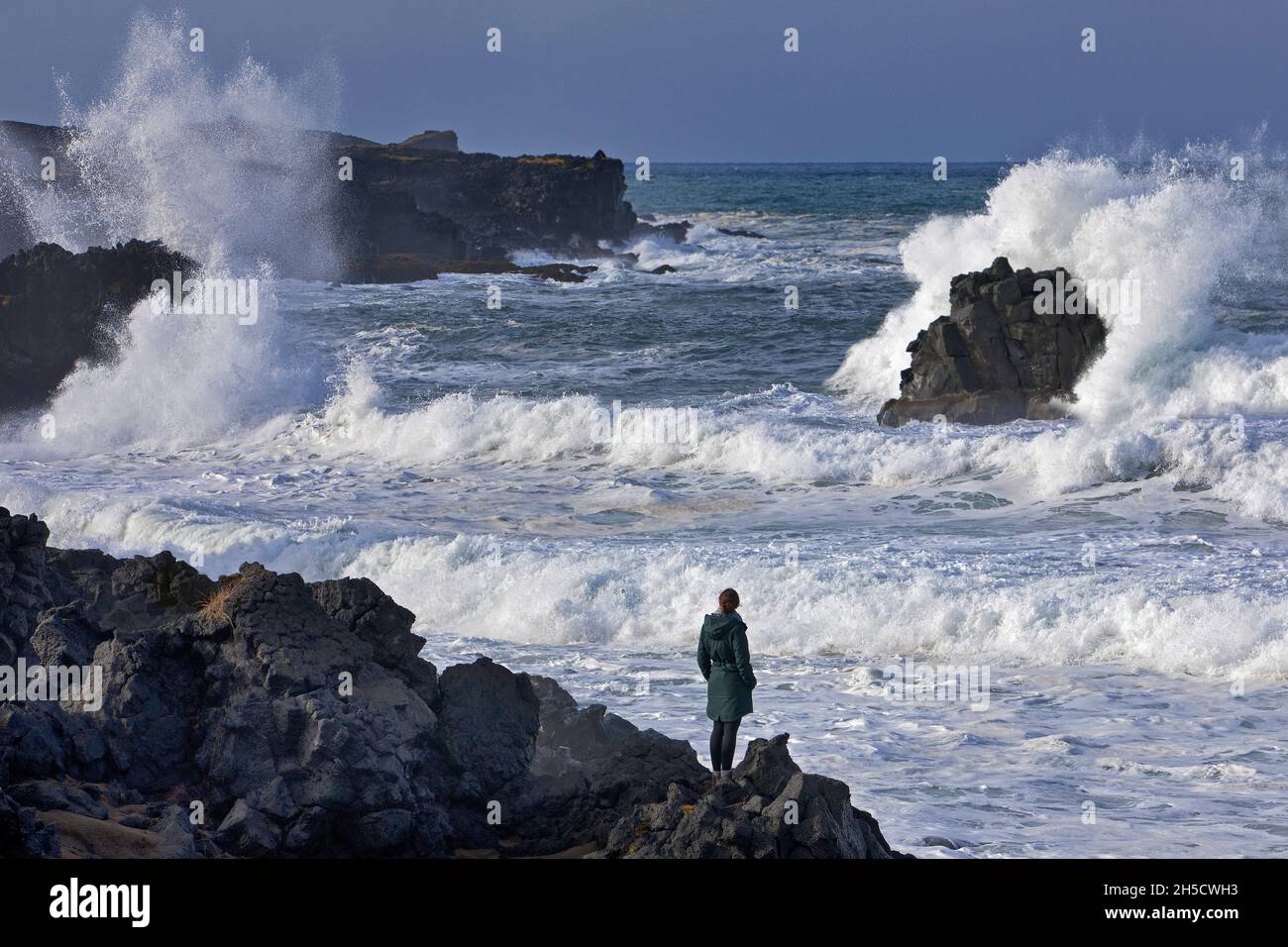 Donna si erge sulla costa di lava nera e guarda il ruggente surf oceano , Islanda, Snaefellsnes, Skardsvik Foto Stock