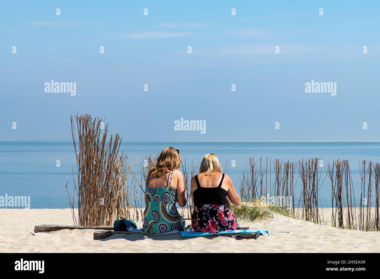 Vista ad angolo basso di due donne visto sulla loro schiena in abito colorato seduto sulla spiaggia dell'isola di Texel, i Paesi Bassi che si affaccia su un mare blu calmo surr Foto Stock