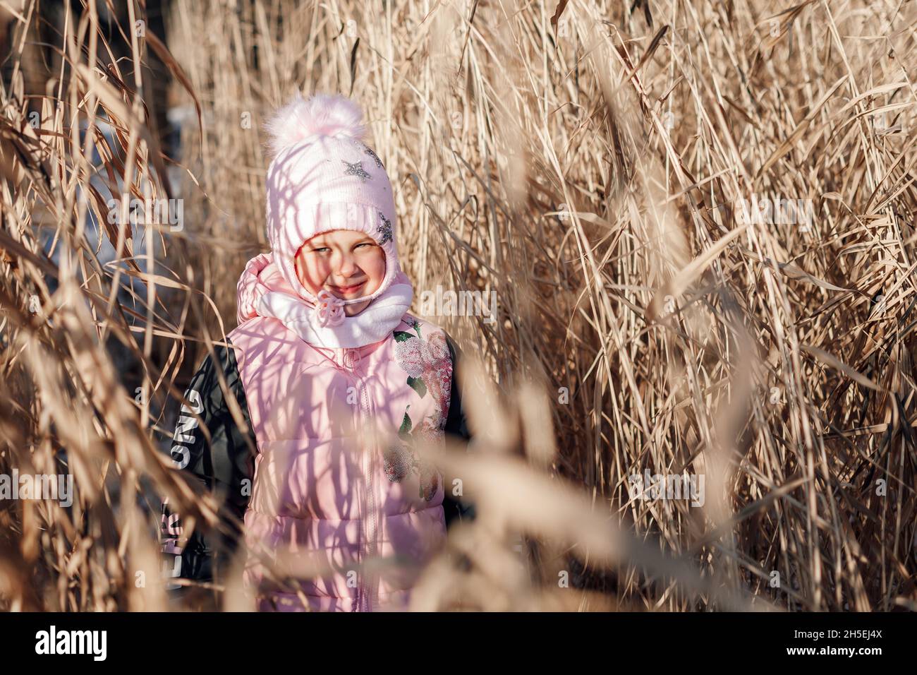 Bambina in erba secca. Bambino in rosa caldo vestito inverno esplora il thicket di canne in giorno di sole nella natura e gode di passeggiata Foto Stock