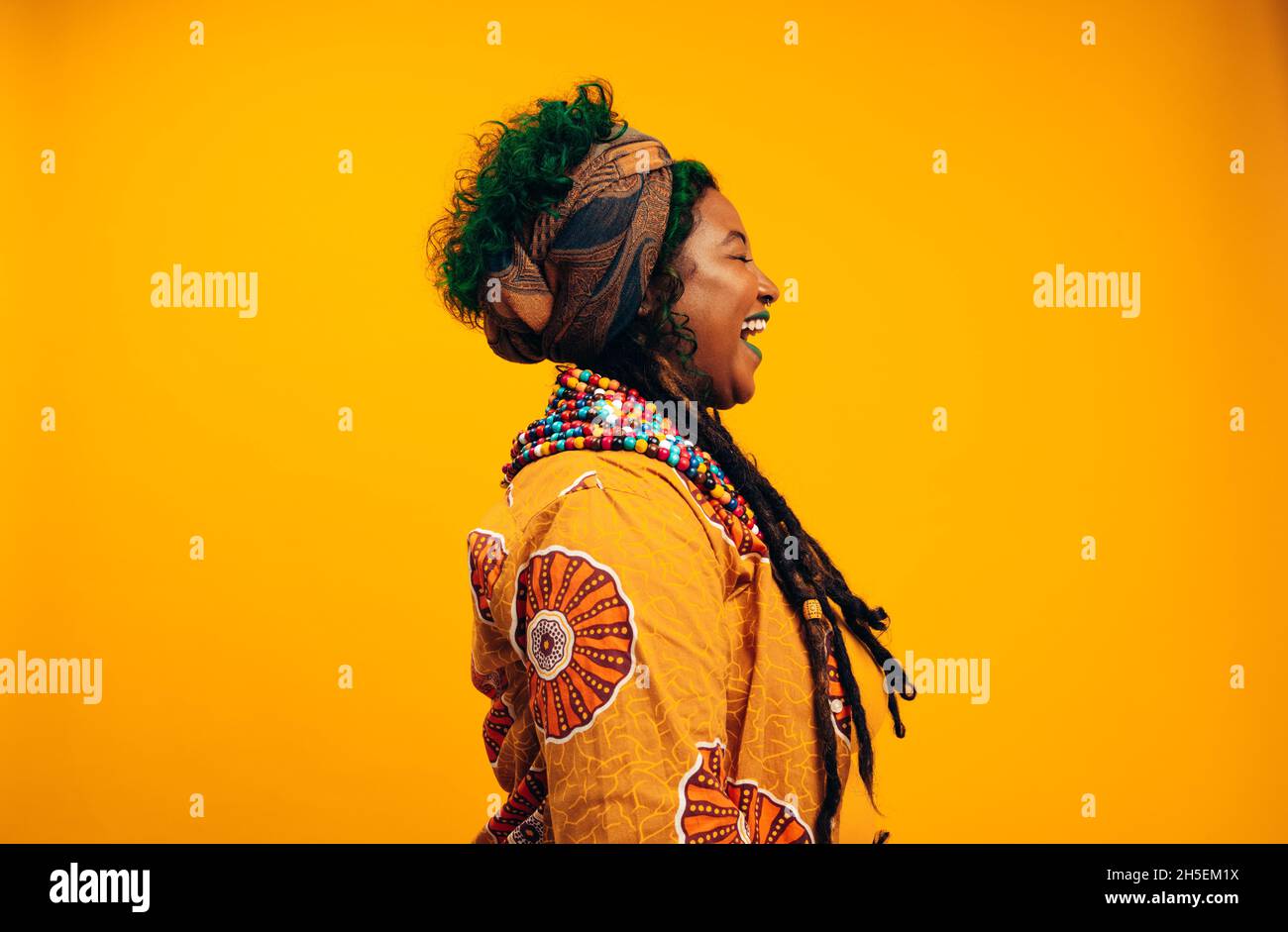 Donna che ride felicemente in uno studio. Giovane donna alla moda che indossa abiti tradizionali africani su sfondo giallo. Abbigliamento elegante da giovane donna Foto Stock