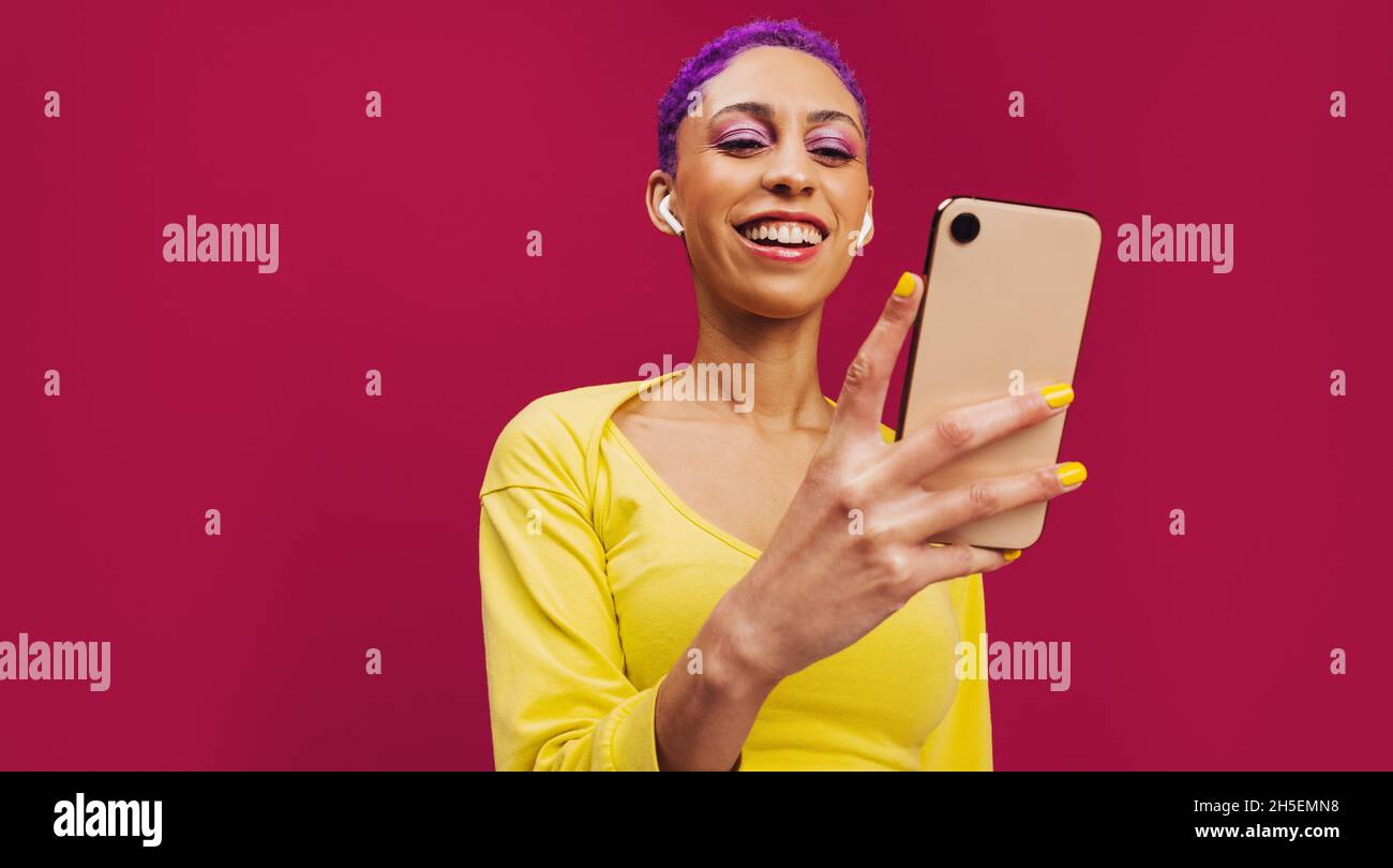 Streaming dei migliori podcast. Giovane donna felice sorridendo allegro mentre tiene uno smartphone e indossa gli auricolari wireless. Donna con capelli viola it Foto Stock