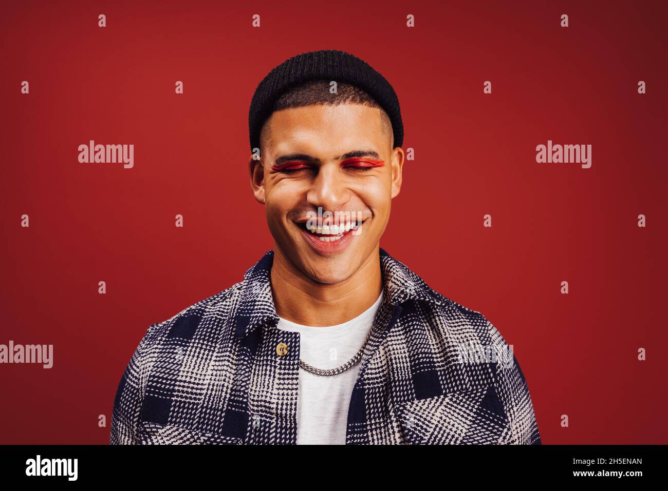 Sensazione grande nel trucco. Giovane uomo felice che indossa l'ombra dell'occhio rosso in uno studio. Giovane uomo alla moda sorridendo con gli occhi chiusi mentre si alza contro Foto Stock