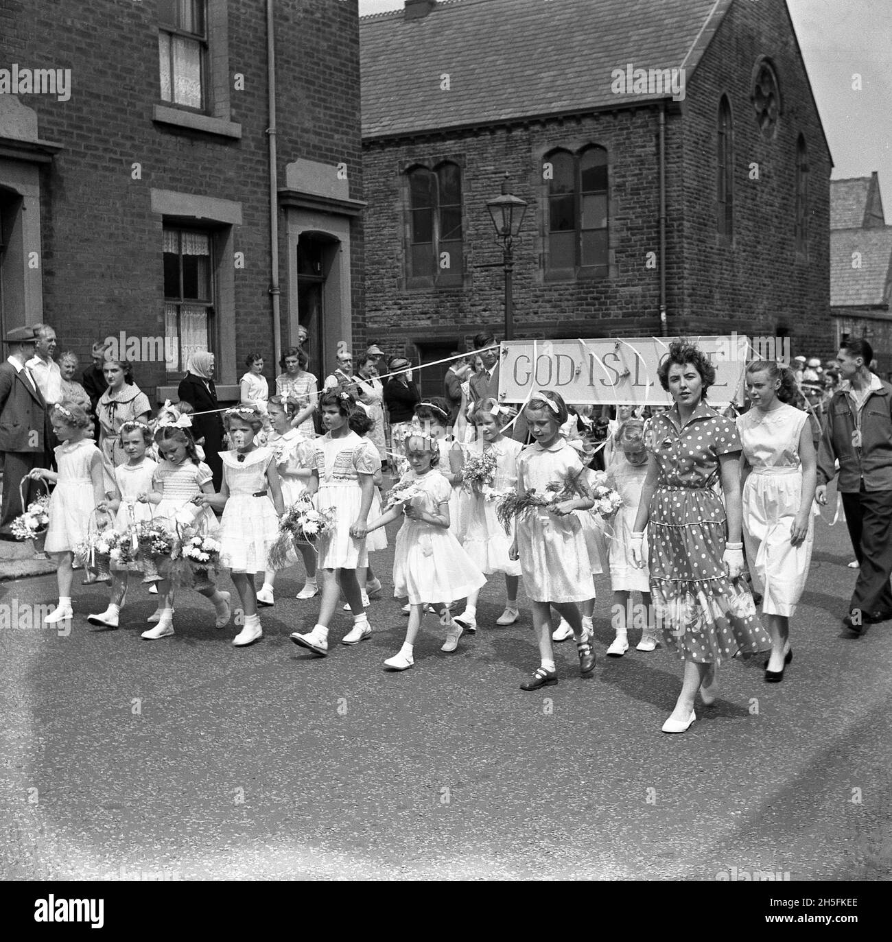 1950, storica, ragazze giovani da una scuola domenicale della Chiesa Congregazionale locale che prendono parte ad una processione di strada nella città di cotone dell'Inghilterra settentrionale di Audley Range, con molte delle ragazze che tengono su una bandiera con le parole, 'Dio è Amore', Audley Range, Blackburn, Lancashire, Inghilterra, Regno Unito. Foto Stock