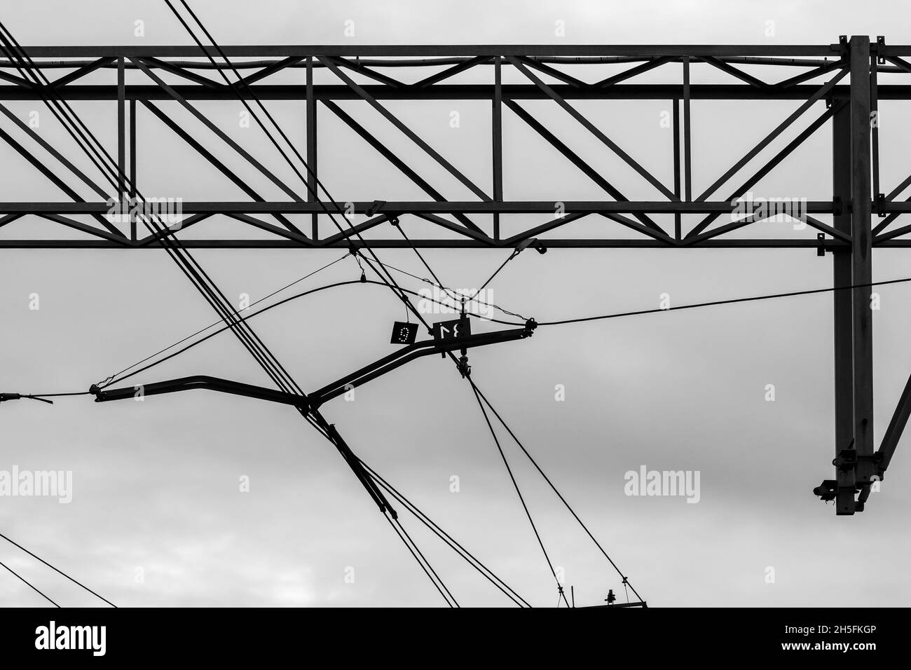 Linee elettriche ferroviarie aeree e strutture di contenimento sotto cielo nuvoloso, foto di sfondo in bianco e nero Foto Stock