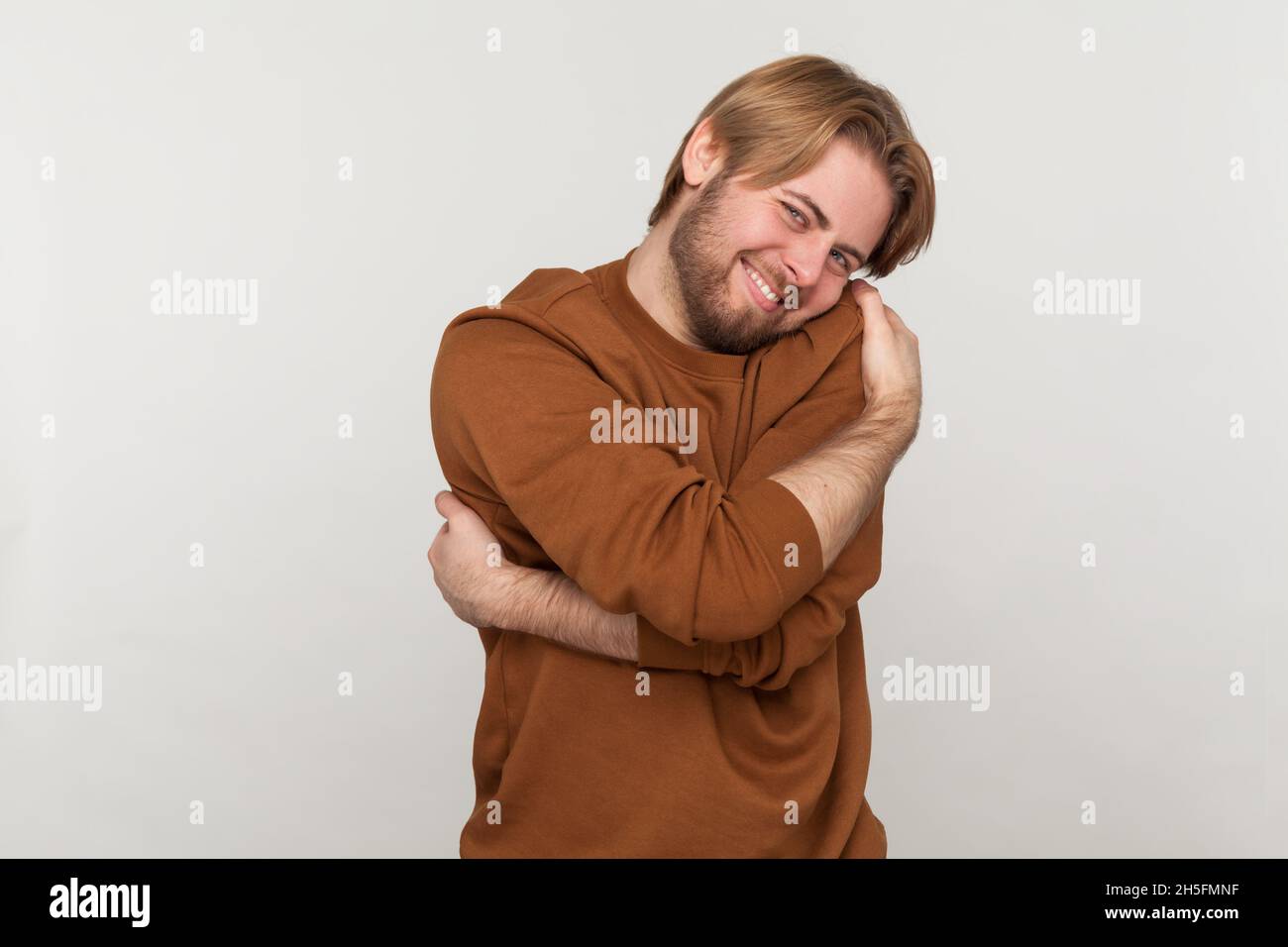 Ritratto di felice bell'uomo con barba che indossa una felpa, in piedi e abbracciandosi con un sorriso toothy godendo, autostima positiva. Studio interno girato isolato su sfondo grigio. Foto Stock