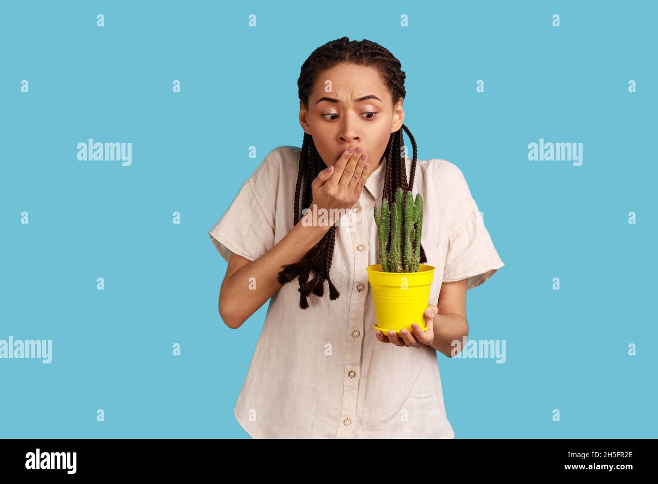 Ritratto di donna spaventata con cialde nere che tengono la pentola del fiore con cactus pungente, guardando la pianta e coprendo la sua bocca, indossando la camicia bianca. Studio interno girato isolato su sfondo blu. Foto Stock