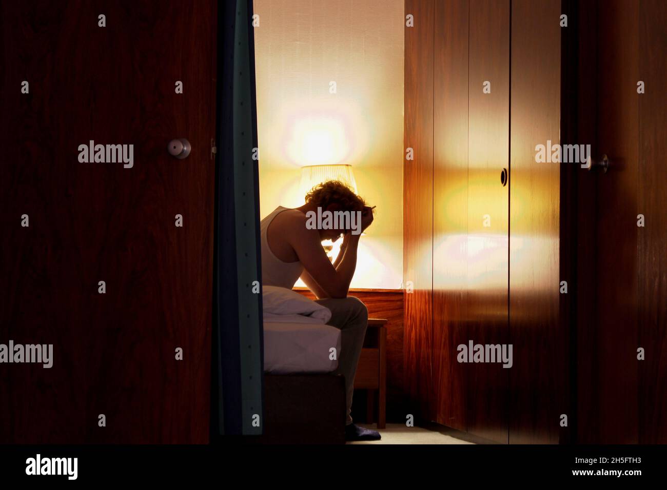 Ein Mann in einem weißen Unterhemd sitzt alleine im Hotelzimmer, den Kopf gesenkt, beide Hände vor der Stirn, mit einer Zigarette in der hand. Foto Stock