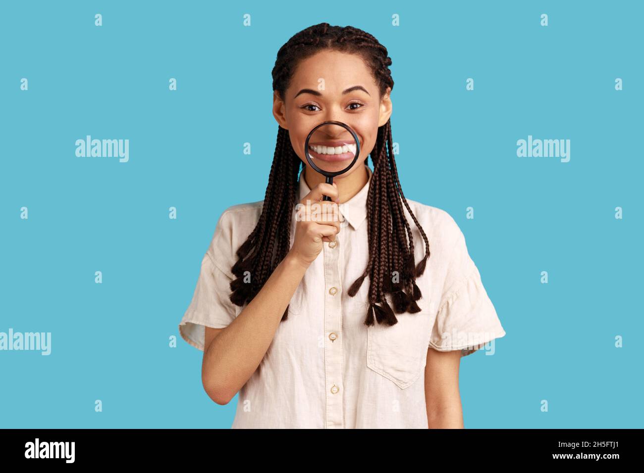 Ritratto di donna sorridente soddisfatto con dreadlock nero che tiene la lente d'ingrandimento sui denti, guardando la macchina fotografica con espressione felice, indossando camicia bianca. Studio interno girato isolato su sfondo blu. Foto Stock