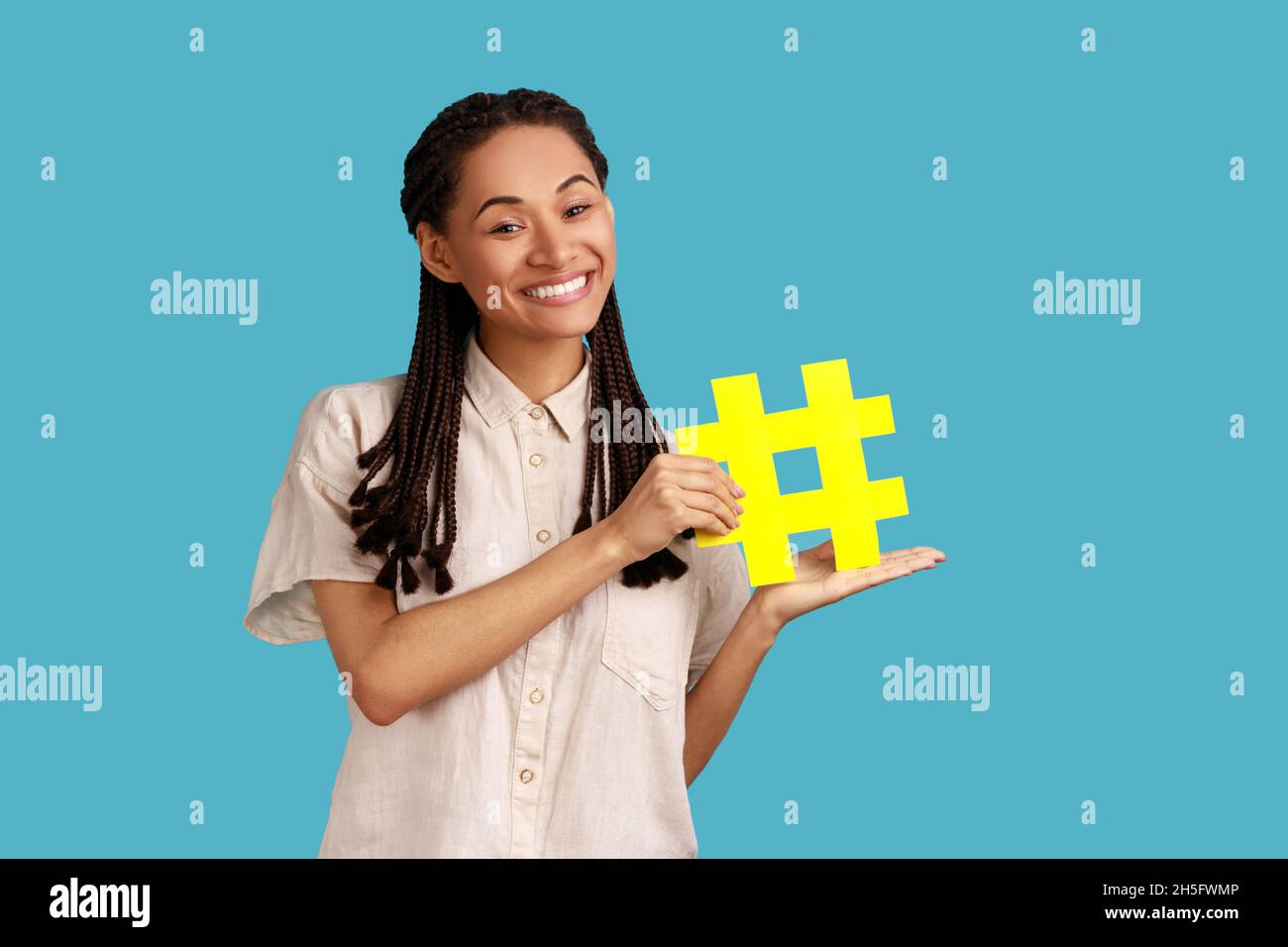 Ritratto di donna felice con nero Dreadlocks mostrando hashtag giallo, comunità internet, famoso contenuto blog, promozione, indossare camicia bianca. Studio interno girato isolato su sfondo blu. Foto Stock