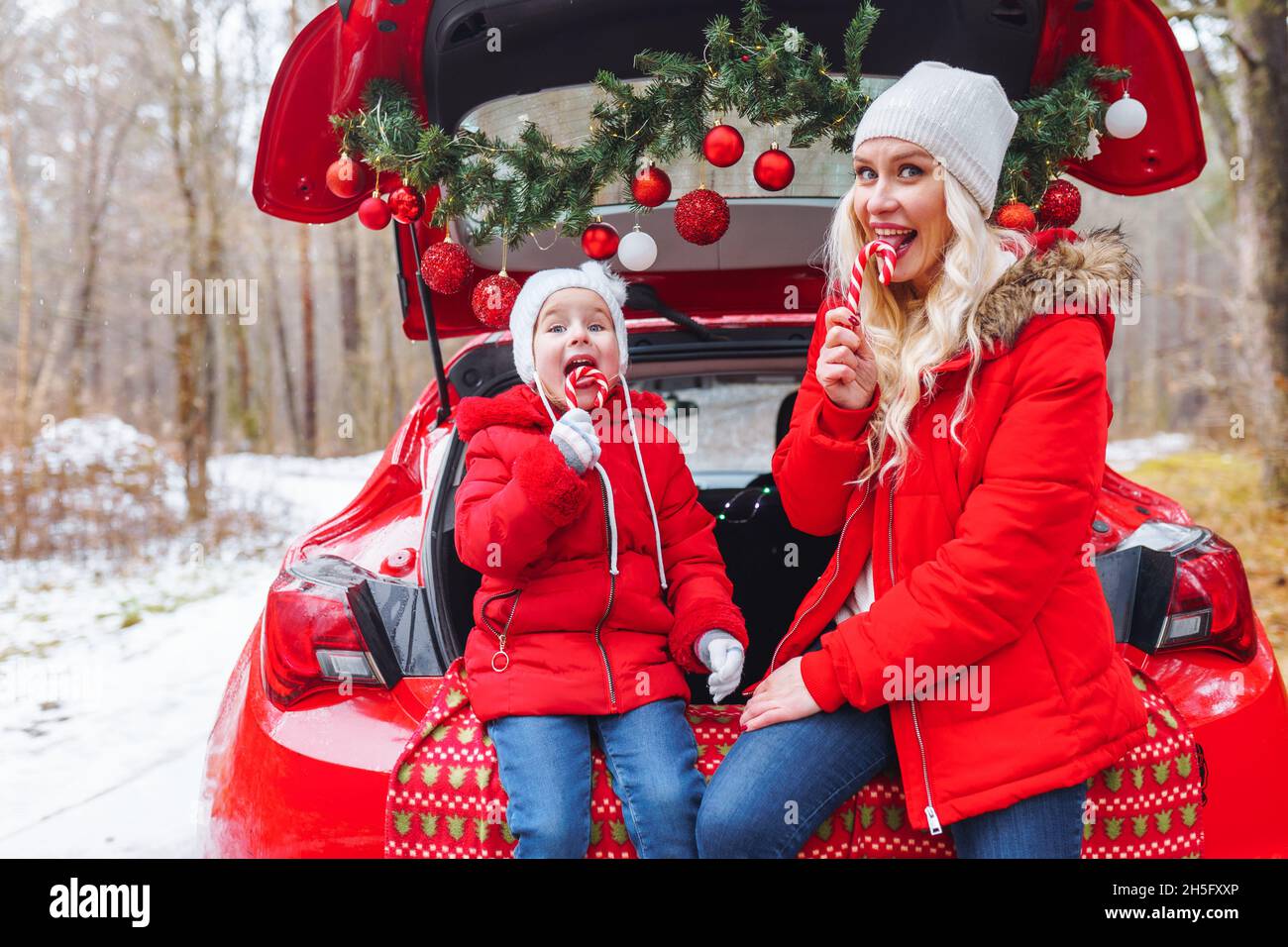 Famiglia felice di mamma bionda e bambina si siedono in auto decorata di natale nella foresta invernale e mangiare caramelle caramelle Foto Stock