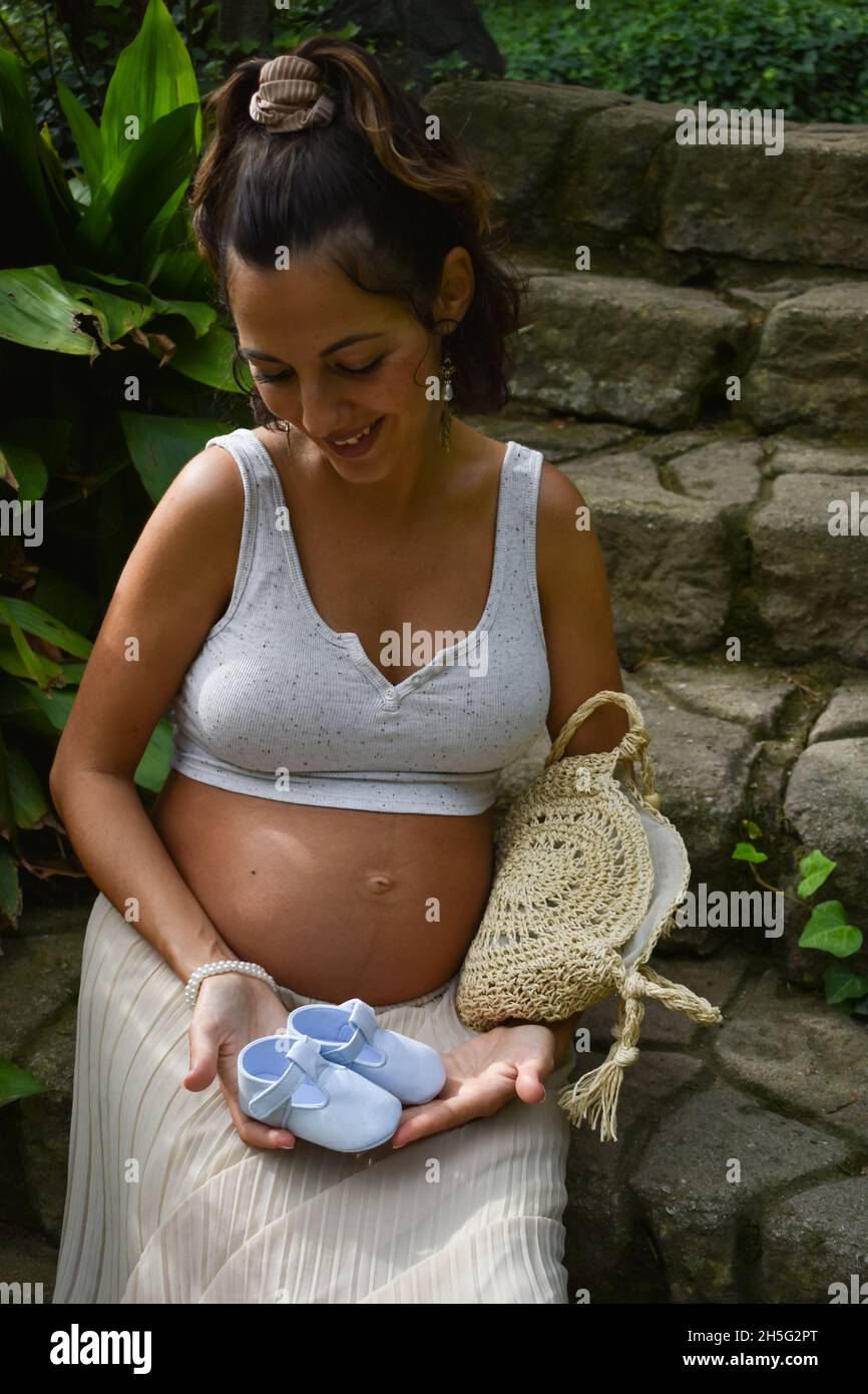 Scatto verticale di una donna incinta caucasica seduta all'aperto e con scarpe da bambino Foto Stock