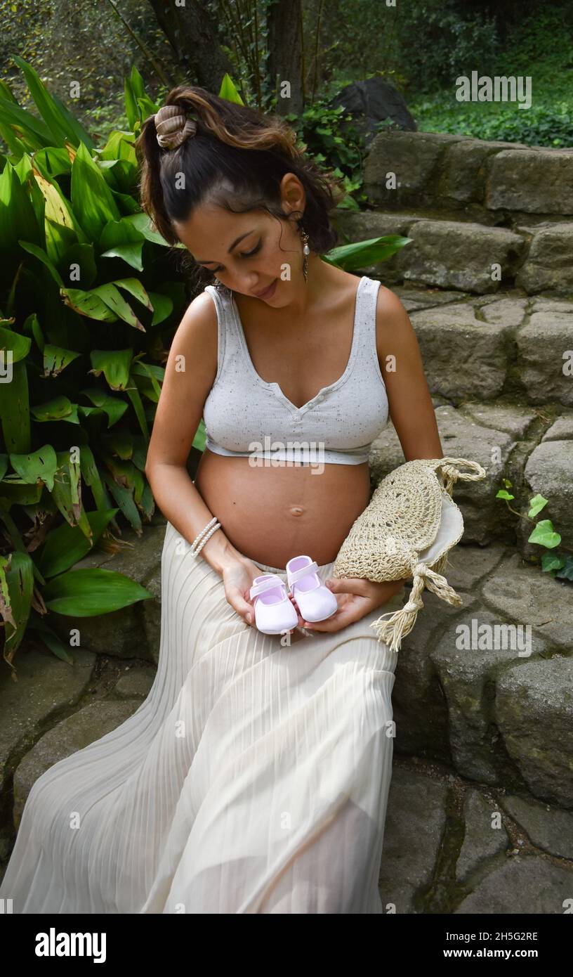 Scatto verticale di una donna incinta caucasica seduta all'aperto e con scarpe da bambino Foto Stock