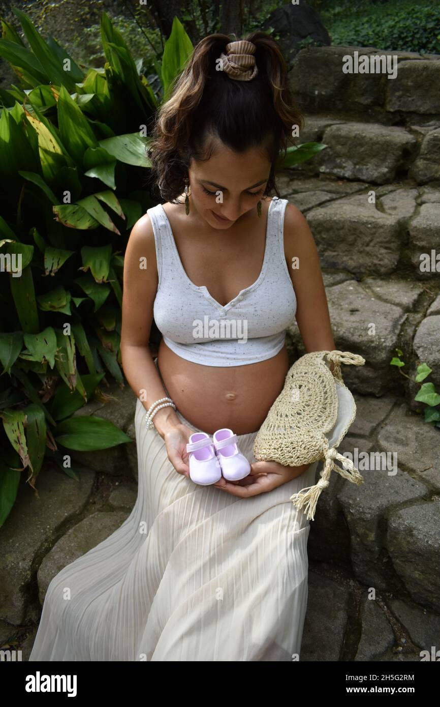 Scatto verticale di una donna incinta caucasica seduta all'aperto e con scarpe da bambino Foto Stock