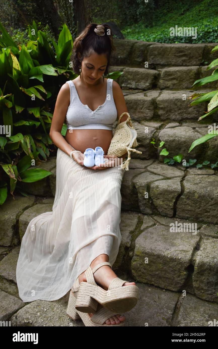 Scatto verticale di una donna incinta caucasica seduta all'aperto e con scarpe da bambino Foto Stock