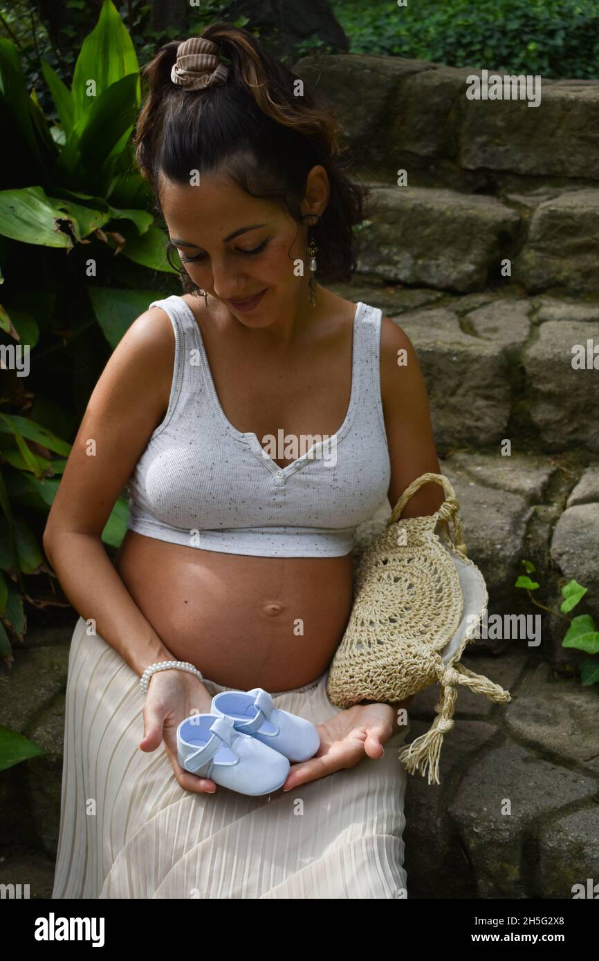 Scatto verticale di una donna incinta caucasica seduta all'aperto e con scarpe da bambino Foto Stock