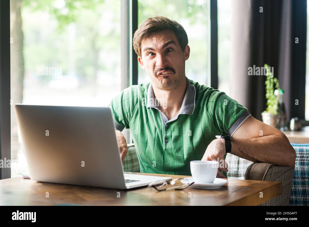Ritratto di un dipendente uomo che indossa una T-shirt verde, che attraversa gli occhi, che si gira intorno e si diverte, seduto sul posto di lavoro di fronte al laptop. Foto al coperto vicino alla grande finestra, sfondo del caffè. Foto Stock