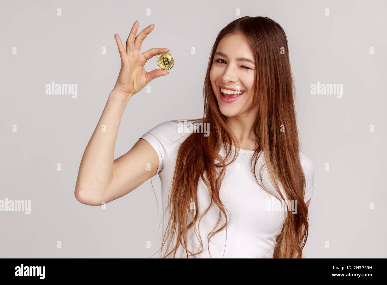 Ritratto di bella donna che tiene il bitcoin d'oro, mostrando la moneta btc e il winking alla macchina fotografica con espressione positiva, indossando la T-shirt bianca. Studio interno girato isolato su sfondo grigio. Foto Stock