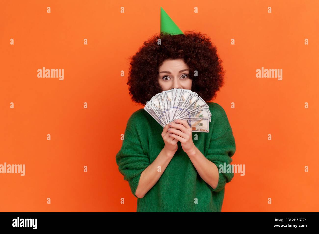 Donna felice con acconciatura afroafroche indossa maglione verde casual stile che tiene coprire metà della bocca con soldi, avendo grande somma di soldi come regalo di compleanno. Studio interno girato isolato su sfondo arancione Foto Stock