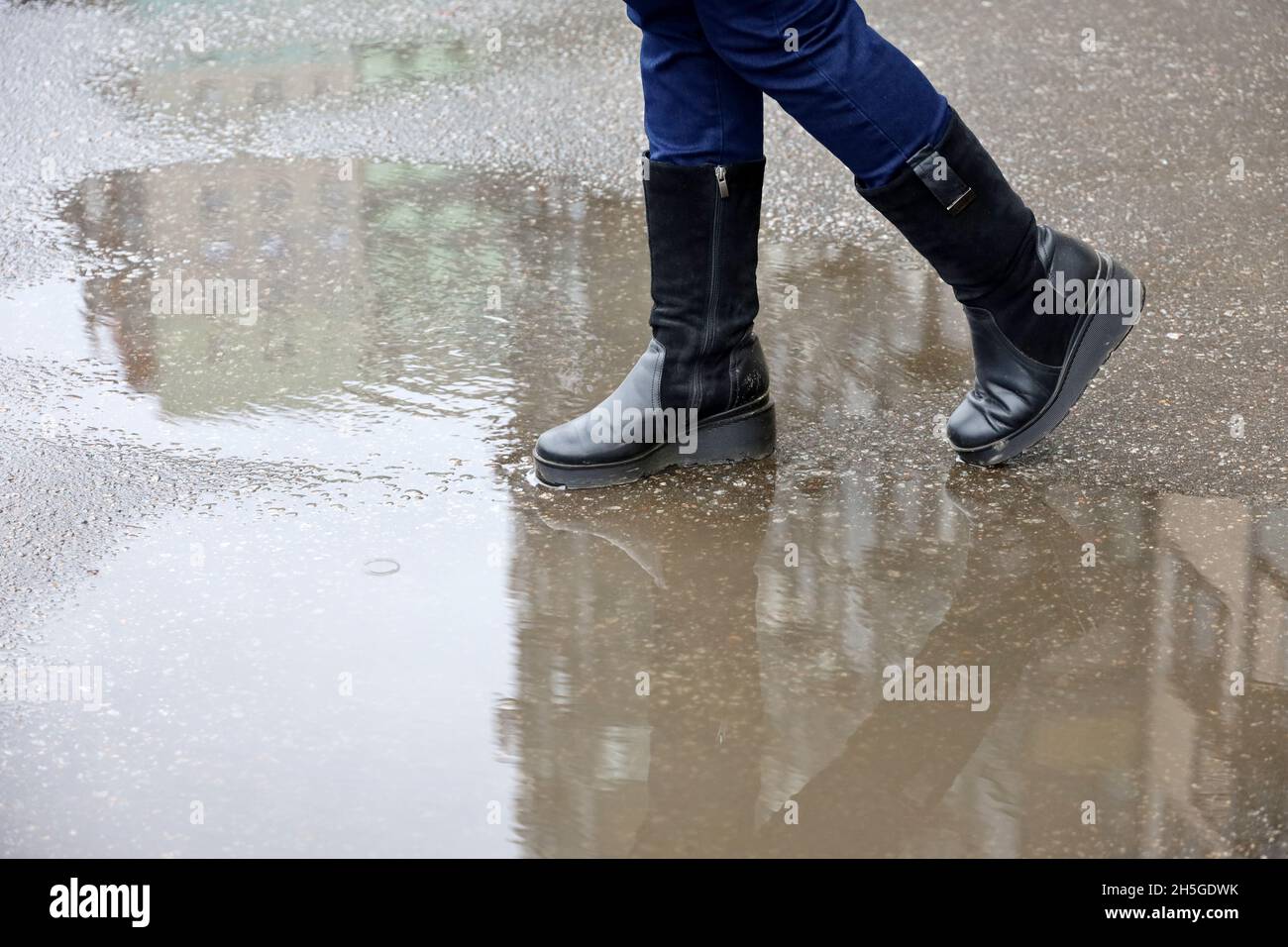 Pioggia in città, le gambe femminili in stivali su una strada con pozzanghere. Gli edifici residenziali si riflettono in acqua, le donne che camminano in autunno tempo Foto Stock