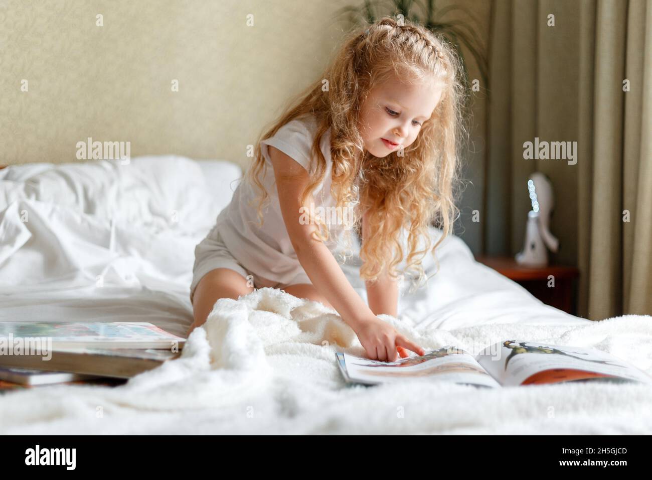 Bella bambina bambino leggere il libro a letto al mattino. Piccola bionda ragazza curly leggere libro in camera da letto soleggiato stanza dei bambini a casa. Ragazza caucasica Foto Stock