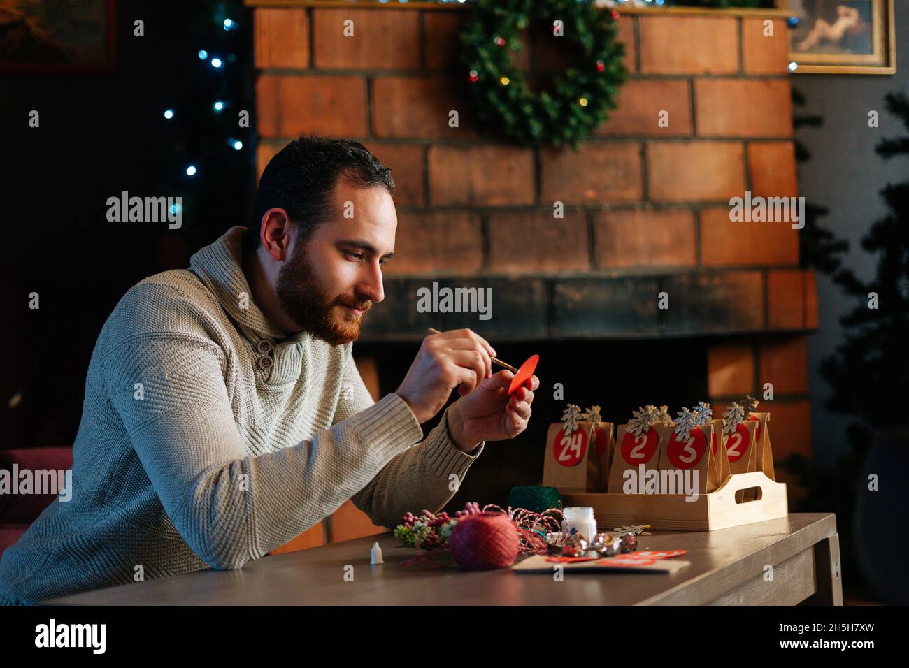 Vista laterale del giovane uomo che scrive numero sul sacchetto rosso che fa i sacchetti di carta da carta di kraft per calendario dell'avvento sulla vigilia di Natale seduta dal camino. Foto Stock