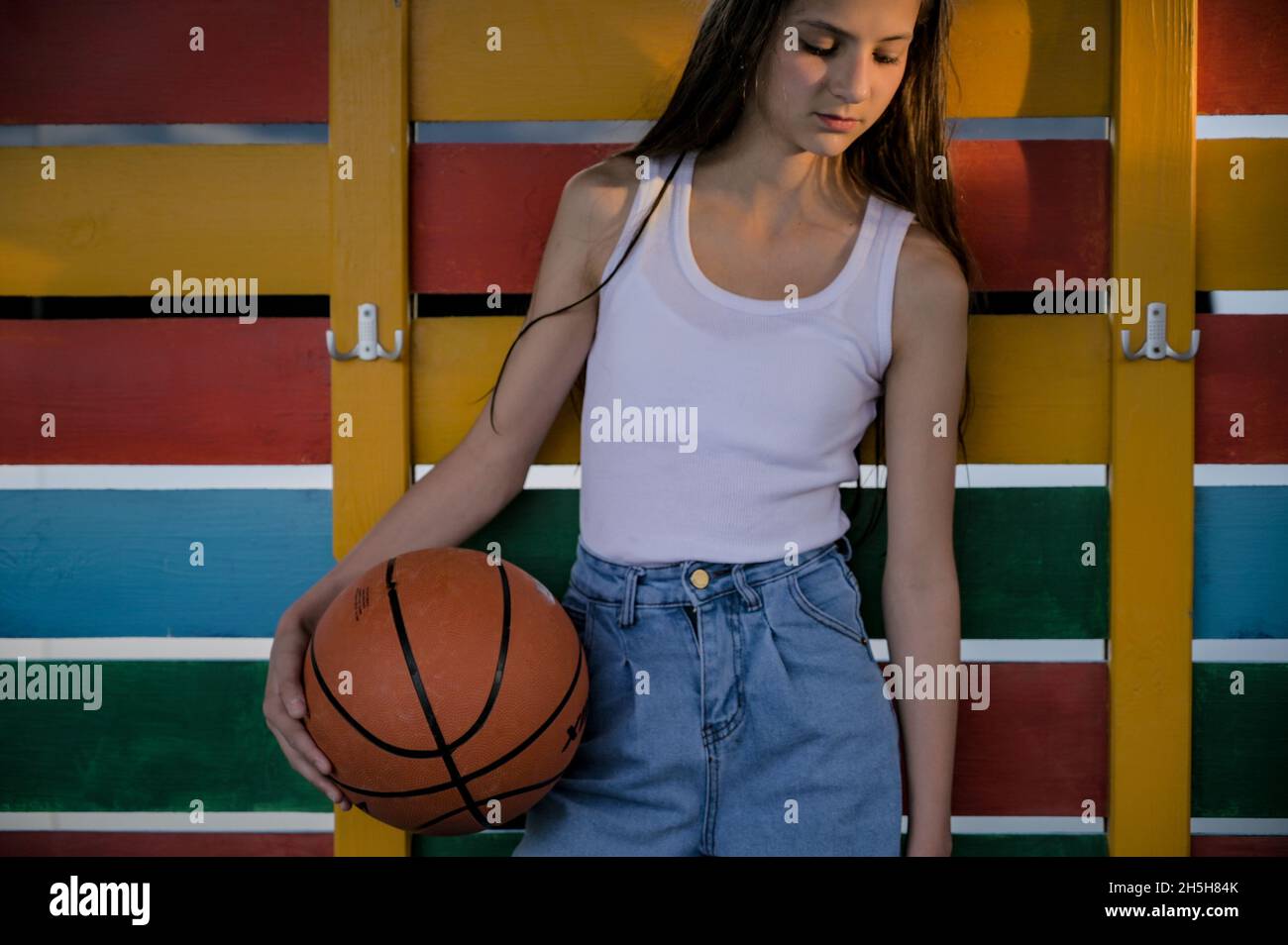bella piccola ragazza giovane in bianco canotta top e jeans blu denim che tiene palla da basket su colore sfondo guardaroba in legno Foto Stock
