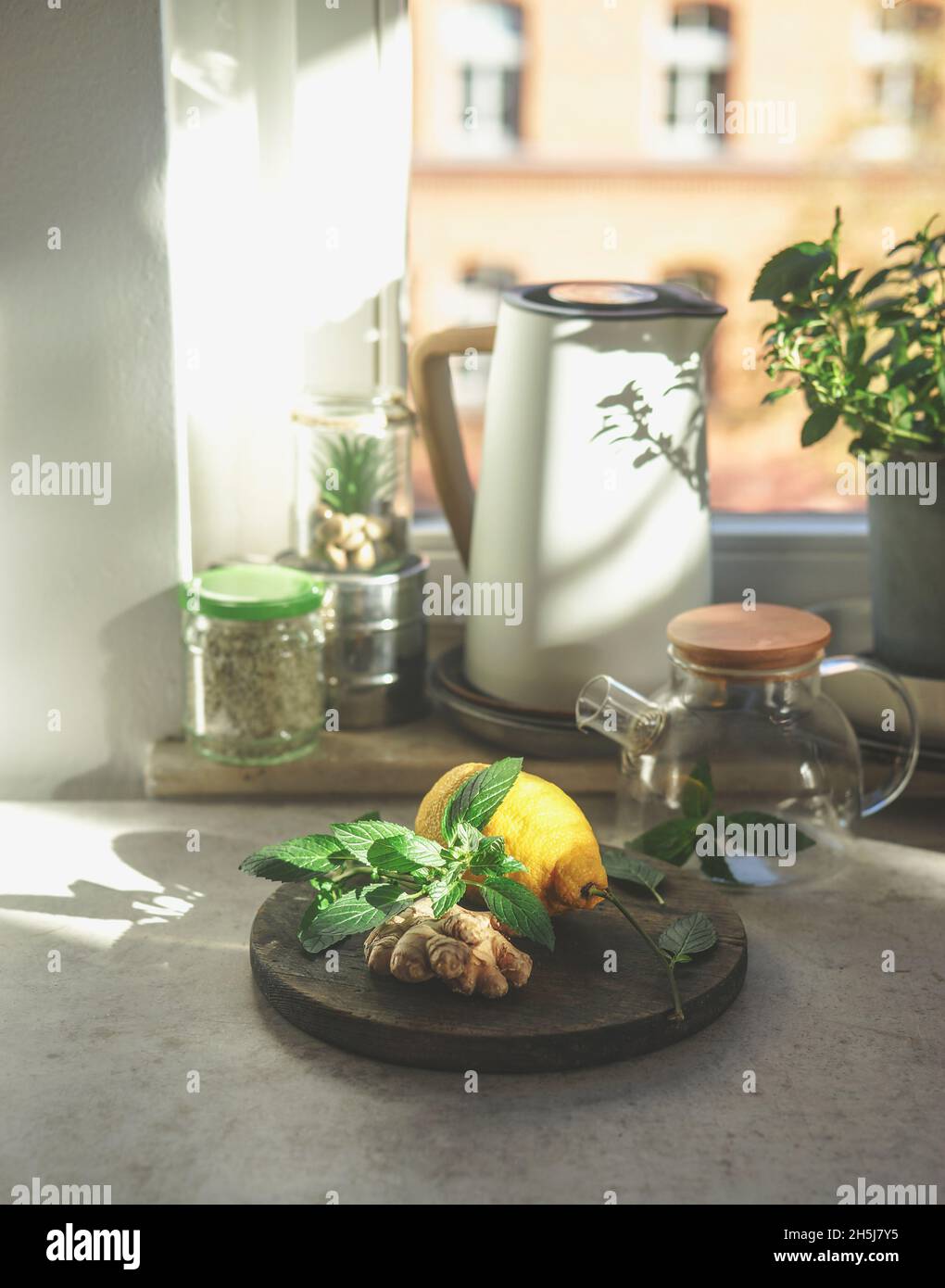 Preparazione di tè allo zenzero fresco con ingredienti sani a casa. Zenzero, foglie di menta e limone sul tavolo da cucina con tea pot in vetro, bollitore elettrico e ja Foto Stock