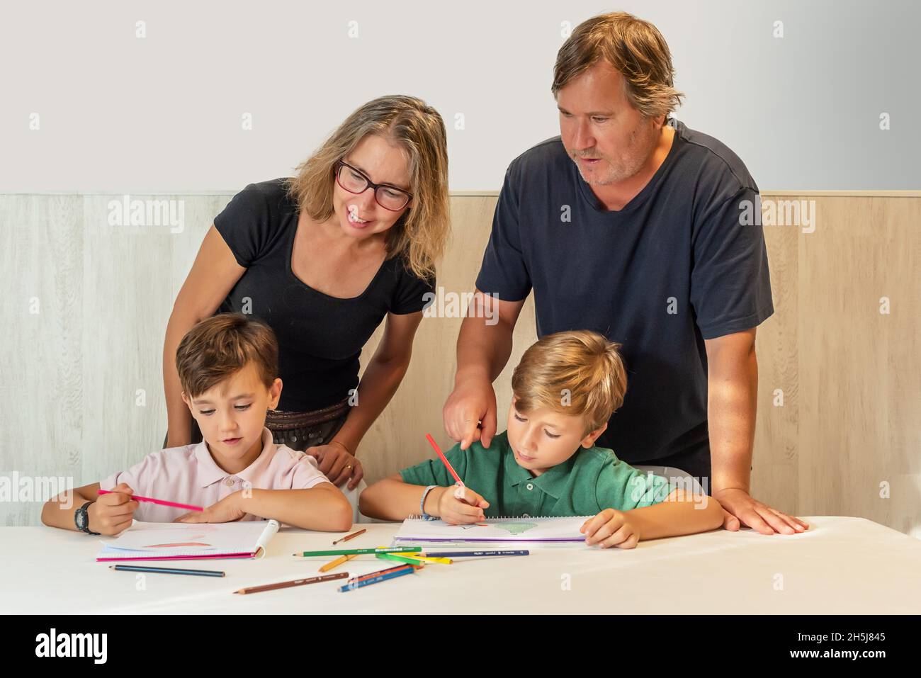 Due bambini fanno i loro compiti a casa mentre i loro genitori li guardano e li assistono Foto Stock
