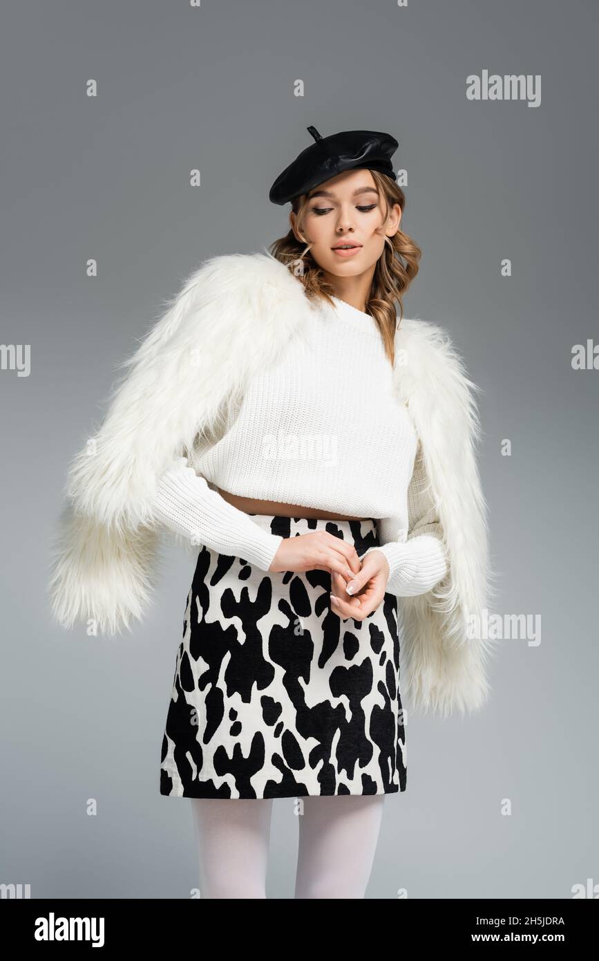 giovane donna in beretta, giacca bianca in pelliccia sintetica e gonna con stampa animale in posa isolata sul grigio Foto Stock