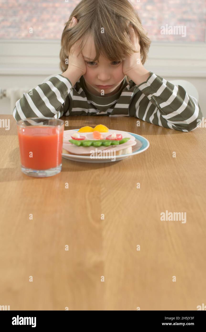 Ragazzo frustrato seduto al tavolo di fronte a un panino sano con verdure e un bicchiere di succo di pomodoro; Germania Foto Stock