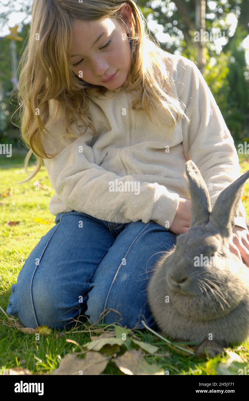 Ragazza che si inginocchia sull'erba accarezzando un coniglio grigio (Oryctolagus cuniculus domesticus); Germania Foto Stock