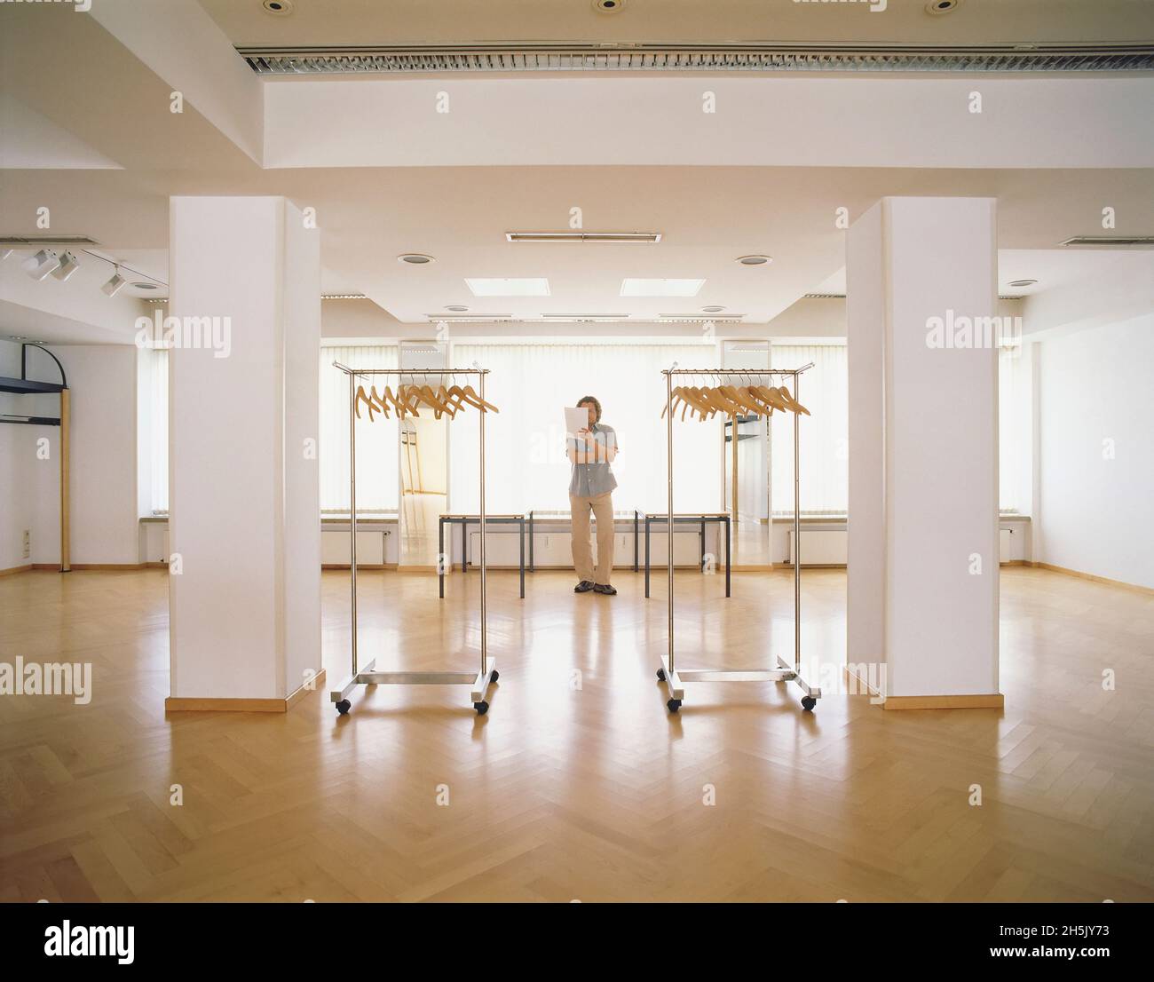 Assistente di vendita maschile che legge un documento mentre si trova in piedi in un negozio con appendiabiti portatili in primo piano; Germania Foto Stock