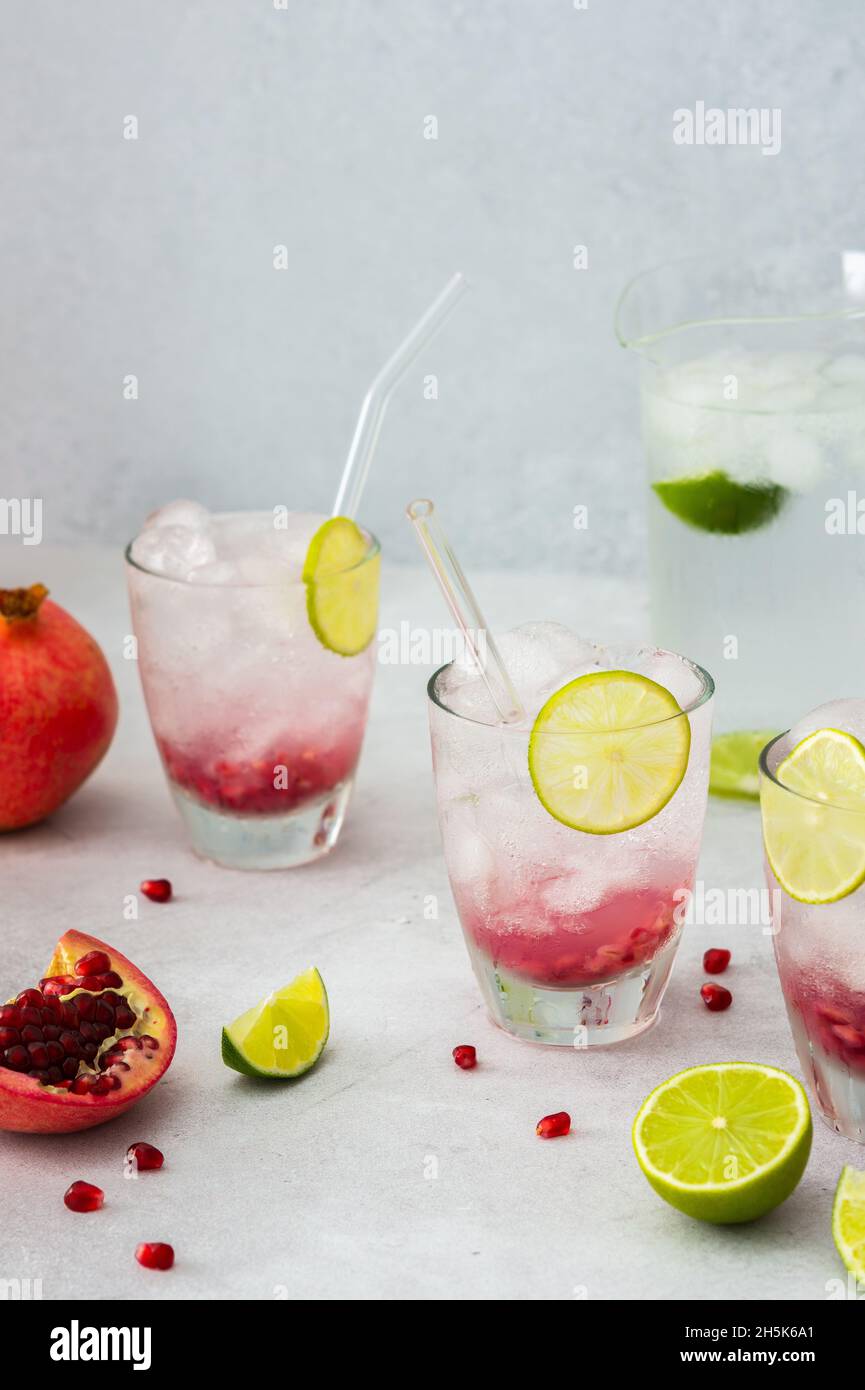 Rinfrescante bevanda di melograno in bicchieri con semi di melograno e ghiaccio, cannucce di calce e vetro, brocca di acqua fredda con agrumi, melograni tagliati Foto Stock